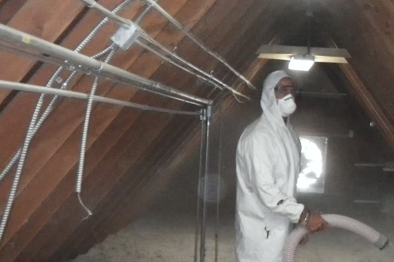 A worker in a white protective suit and dust mask blows loose-fill (blown-in) insulation through a hose across an attic floor.