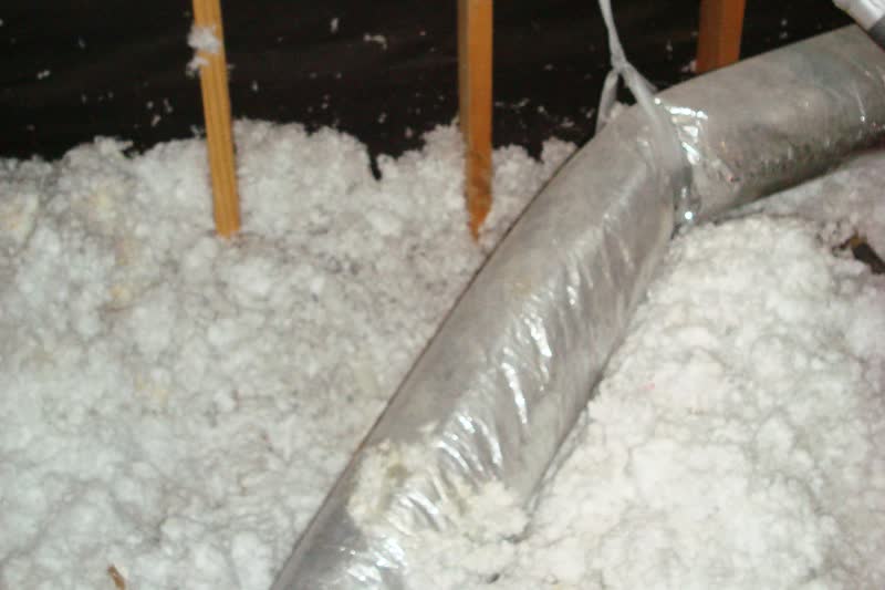 An insulated flexible air duct with a silver jacket lying across white loose-fill insulation in an attic.