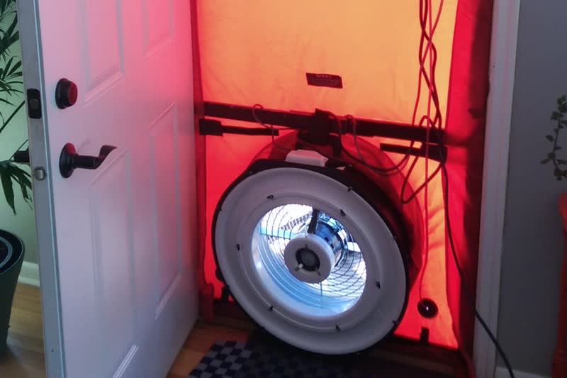 A blower door test set up in a front doorway: a red fabric panel seals the opening around a large round fan.