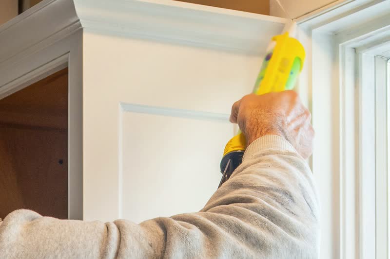 A hand guides a caulk gun along the trim at the top of a window frame.