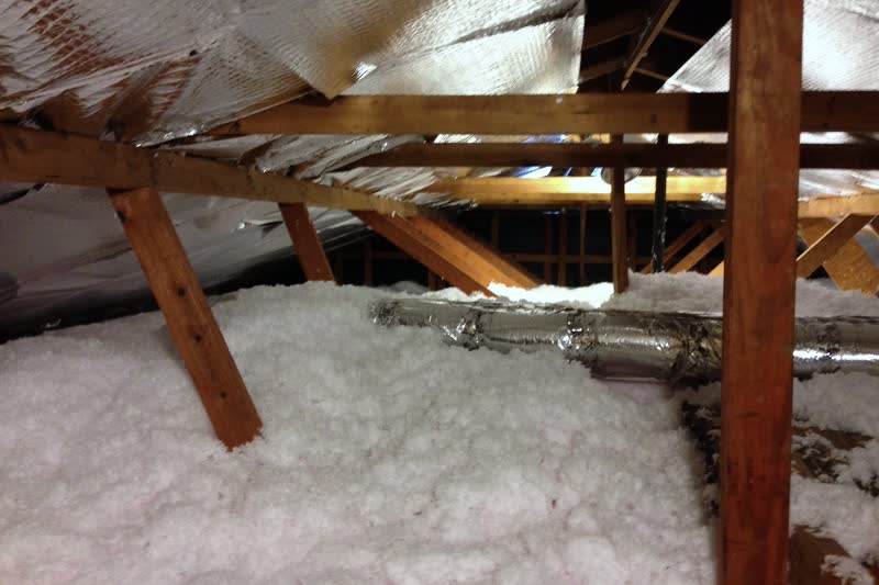 White blown-in (loose-fill) insulation covering an attic floor around roof truss webs, with a foil-wrapped flexible duct lying on top.