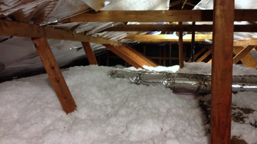 White blown-in (loose-fill) insulation covering an attic floor around roof truss webs, with a foil-wrapped flexible duct lying on top.