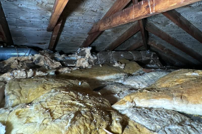Old, uneven yellow fiberglass batt insulation covering an attic floor under a plywood roof deck.