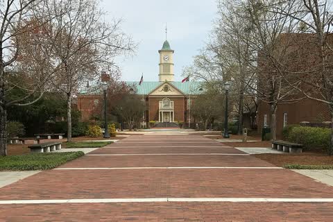 Brick-paved promenade between bare trees leading to a red-brick building with a white clock tower and two flags