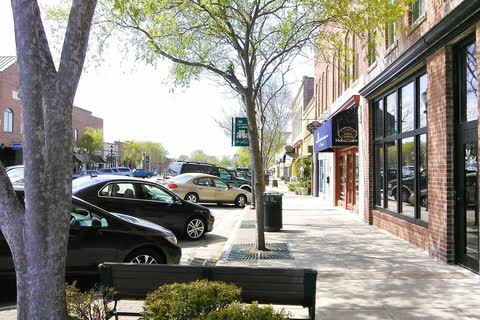 A tree-lined sidewalk beside brick storefronts in downtown Fuquay-Varina