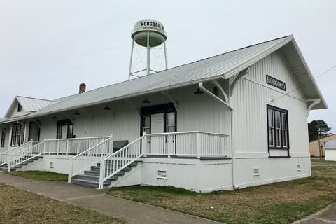 A restored white wooden railroad depot with the Hobgood water tower behind