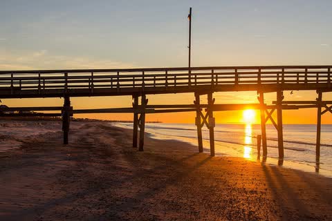 The sun rising behind the pilings of a long fishing pier on the beach at Oak Island