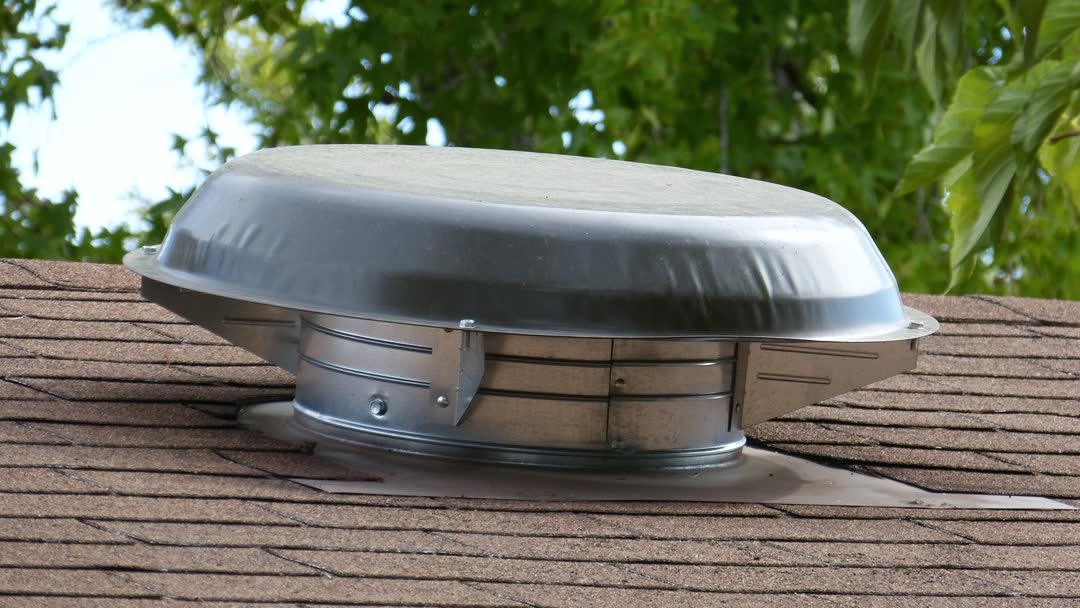 A powered attic fan with a round metal cap mounted on an asphalt shingle roof, with trees behind it.