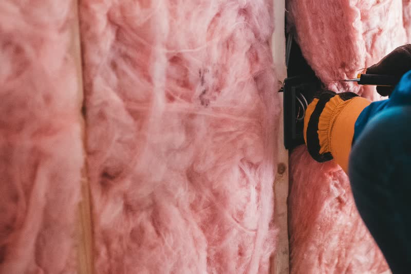 Pink fiberglass batt insulation fitted between the wood studs of an open wall, with a gloved hand at an electrical box.